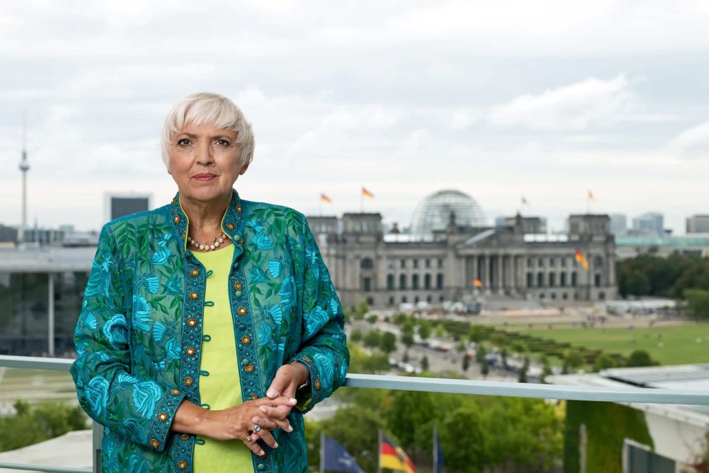 Claudia Roth steht in einer bestickten, blau-grünen Jacke und hellgrünem Oberteil auf einem Balkon und blickt in die Kamera. Im Hintergrund sieht man die Kuppel des Reichstags und den Berliner Fernsehturm.