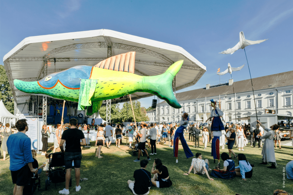 Sommerliches Fest im Park von Schloss Bellevue mit vielen Besucherinnen und Besuchern, bunter Riesenfischfigur vor einer Bühne, Stelzenläuferin und -läufer und Dekorationen in Form von weißen Vögeln.