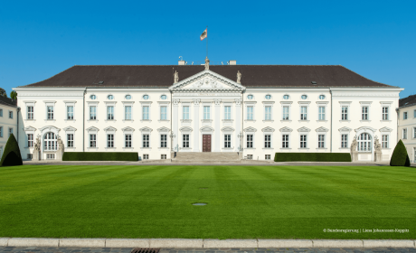 Frontansicht des Berliner Schlosses Bellevue bei blauem Himmel, mit gepflegtem Rasen im Vordergrund und einer Flagge mit dem Bundeswappen auf dem Dach.