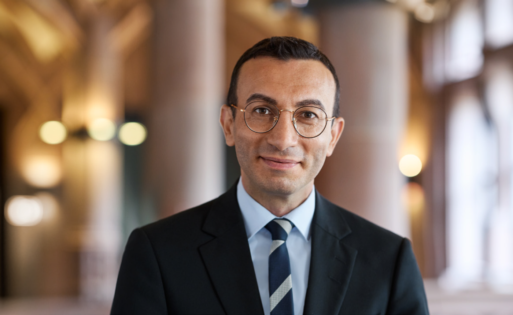 Oberbürgermeister Mike Josef lächelt in die Kamera. Er trägt einen dunklen Anzug. Im Hintergrund sind unscharf Säulen und Lichter zu sehen.