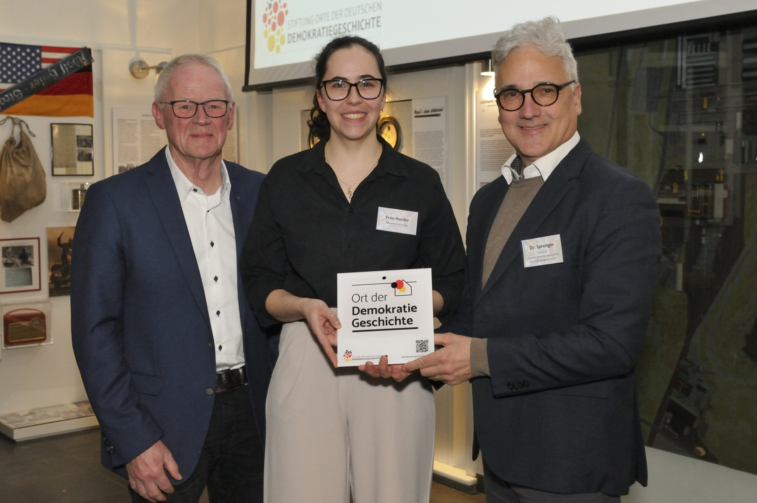 Drei Personen, links ein Mann im dunklen Anzug mit weißen Hemd, mittig eine Frau in schwarzer Bluse, rechts ein Mann inHemd, Pullover und Sakko. Die Personen rechts halten gemeinsam eine weiße, quadratische Acrylplakette, auf der "Ort der Demokratiegeschichte" steht.
