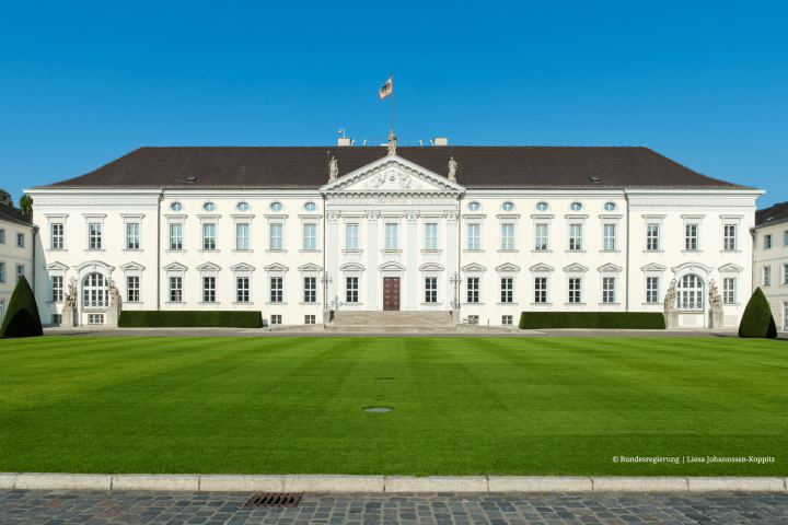 Frontansicht des Berliner Schlosses Bellevue bei blauem Himmel, mit gepflegtem Rasen im Vordergrund und einer Flagge mit dem Bundeswappen auf dem Dach.