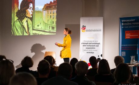 Eine Schauspielerin in gelber Jacke steht nach links gewandt vor Publikum auf einer Bühne. Sie verkörpert das Grundgesetz. An der Wand sieht man eine Präsentation. Rechts auf der Bühne stehen zwei Roll-Ups.