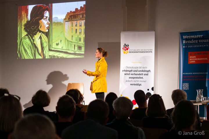 Eine Schauspielerin in gelber Jacke steht nach links gewandt vor Publikum auf einer Bühne. Sie verkörpert das Grundgesetz. An der Wand sieht man eine Präsentation. Rechts auf der Bühne stehen zwei Roll-Ups.