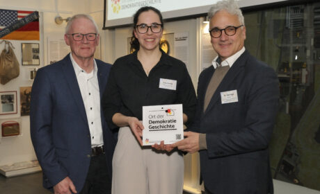 Drei Personen, links ein Mann im dunklen Anzug mit weißen Hemd, mittig eine Frau in schwarzer Bluse, rechts ein Mann inHemd, Pullover und Sakko. Die Personen rechts halten gemeinsam eine weiße, quadratische Acrylplakette, auf der "Ort der Demokratiegeschichte" steht.